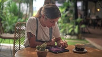 Woman with a ponytail and arm tattoos works on a digital tablet with a stylus, seated at a wooden table with a latte and a cactus.
