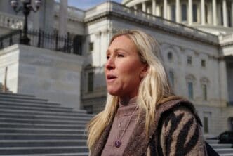 Profile close-up of Rep Marjorie Taylor Greene with long blonde hair, speaking outside the US Capitol Building on a clear day.
