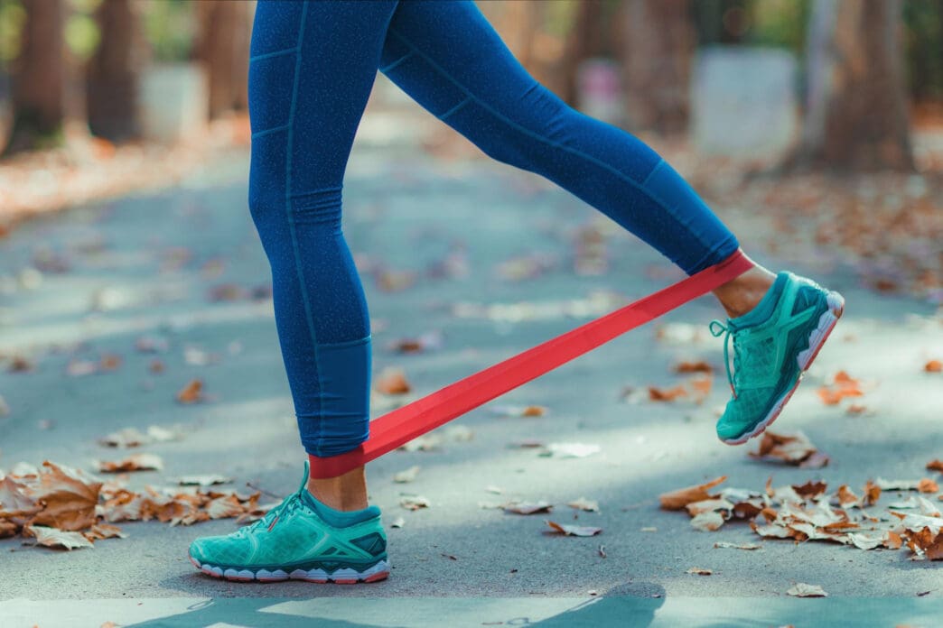 A person is performing a leg exercise with a red resistance band wrapped around their ankles on a leafy outdoor path.