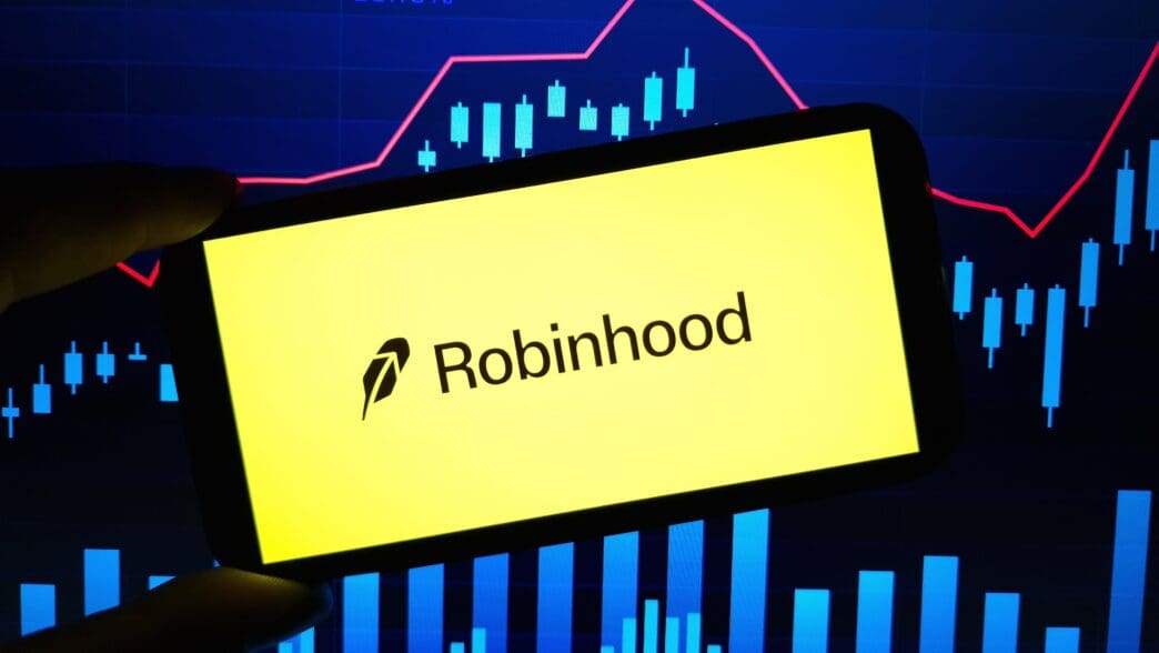 A smartphone displaying the Robinhood logo is held in front of a digital financial graph showing stock market trends.