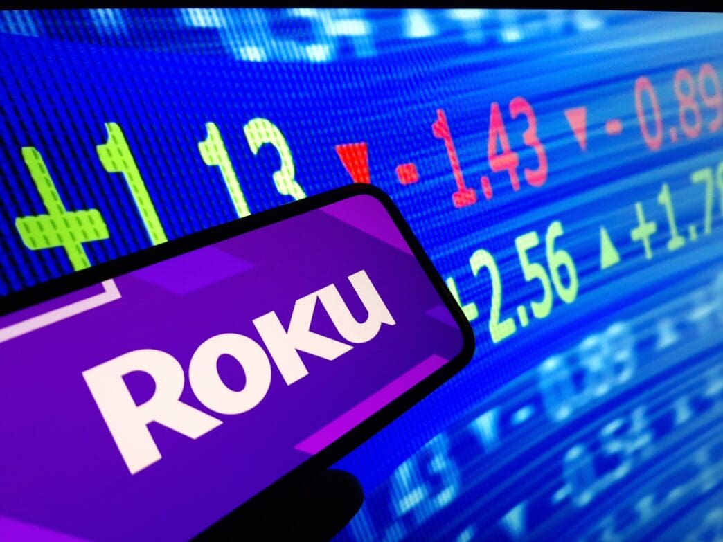 The purple Roku company logo is displayed on a smartphone screen held in front of a blue background showing digital stock market ticker data.