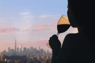 A silhouetted woman sips red wine from a glass while overlooking the New York City skyline from a rooftop at sunset.