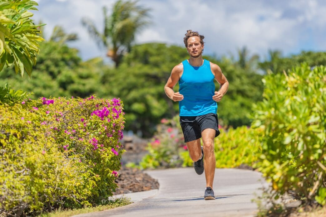 A fit man in a blue tank top and black shorts jogging on a sunny, curved tropical path surrounded by lush greenery