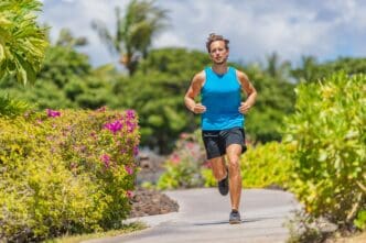 A fit man in a blue tank top and black shorts jogging on a sunny, curved tropical path surrounded by lush greenery