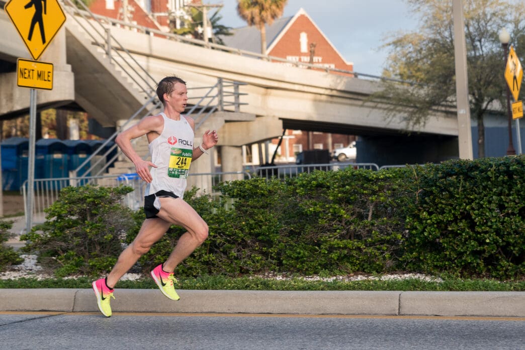 Profile view of a determined male runner wearing a white singlet and bright neon shoes competing in a road race.