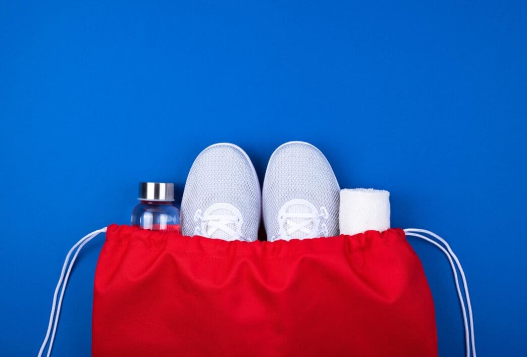 Top-down view of a red drawstring bag containing white athletic shoes, a clear water bottle, and a rolled white towel on a blue background.