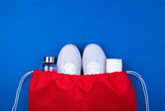 Top-down view of a red drawstring bag containing white athletic shoes, a clear water bottle, and a rolled white towel on a blue background.