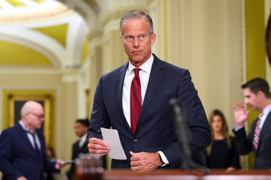 Senate Majority Leader John Thune, dressed in a suit, stands holding a paper inside the U.S. Capitol.