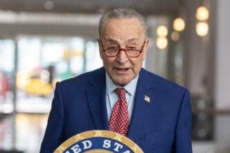 U.S. Senator Chuck Schumer speaks at a press conference, wearing a blue suit, red tie, and red-rimmed glasses, with a seal visible below him.