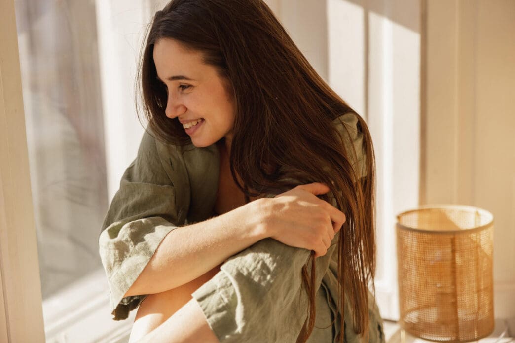 A woman with long brown hair smiles gently while embracing herself by a sunlit window