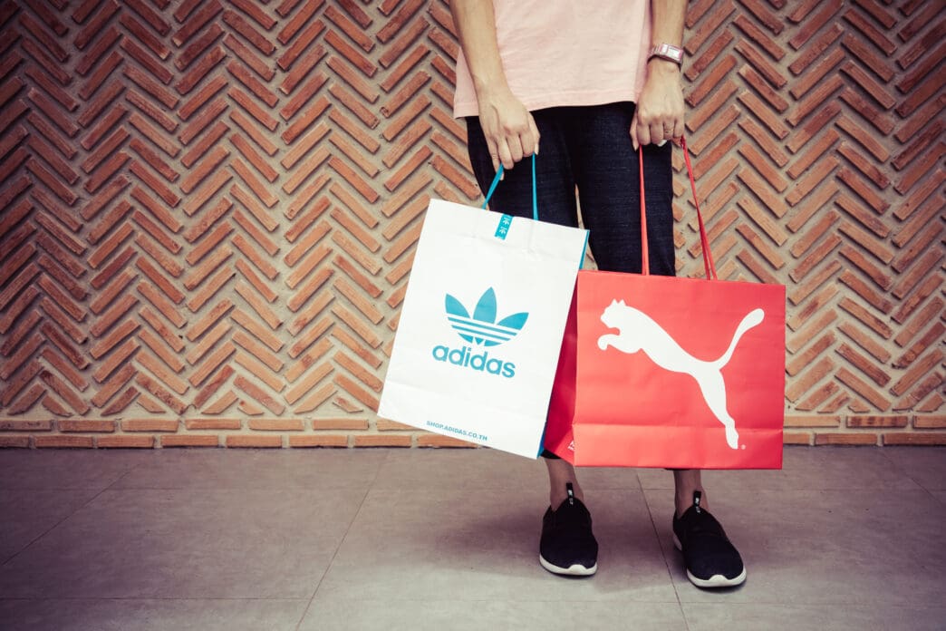 A person holding Adidas and Puma shopping bags in front of a red brick wall with a herringbone pattern