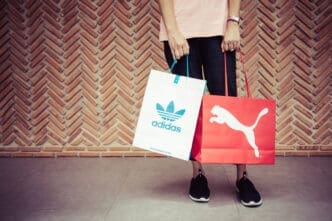 A person holding Adidas and Puma shopping bags in front of a red brick wall with a herringbone pattern