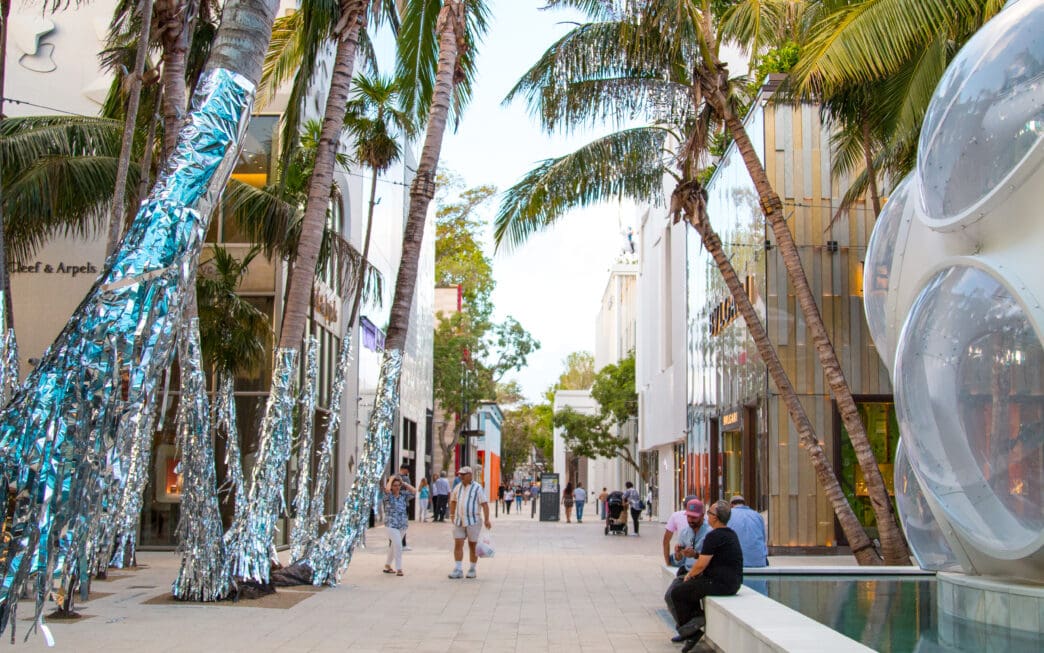 Pedestrians walk down a luxury shopping street lined with palm trees, some wrapped in shiny silver material, in the Miami Design District.
