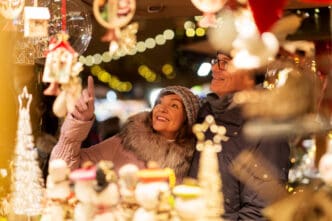 A smiling couple in winter attire looks at holiday decorations in a brightly lit stall at a Christmas market.