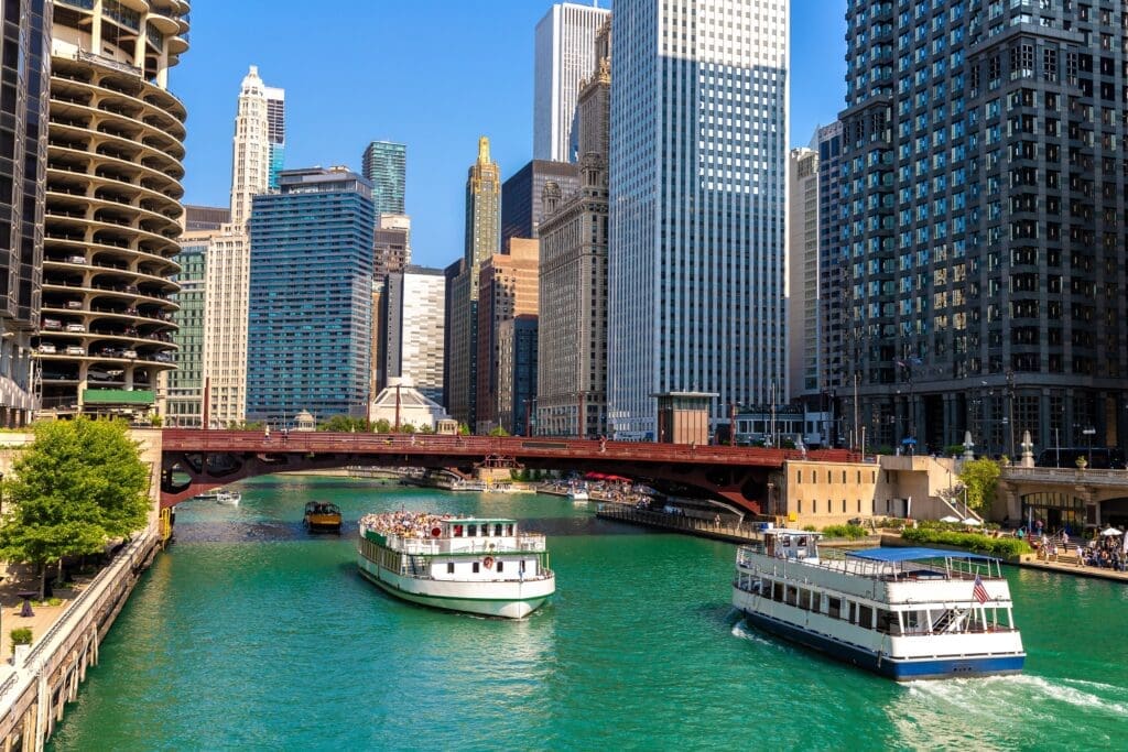 Two sightseeing cruise boats sail on the teal Chicago River, surrounded by skyscrapers.