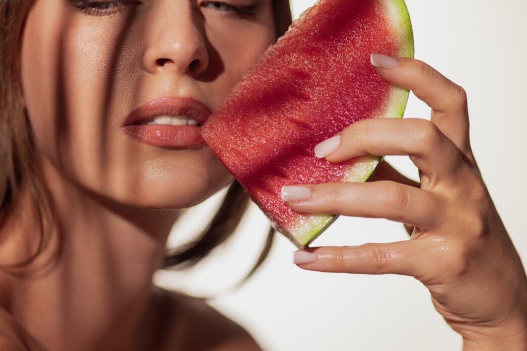Close-up of a woman with wet skin holding a slice of bright red watermelon against her cheek, with shadows across her face