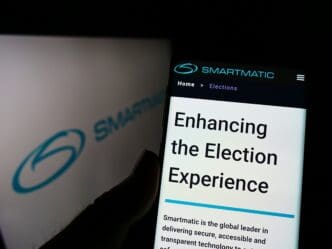 A hand holds a smartphone displaying the Smartmatic website homepage, "Enhancing the Election Experience," with the company logo on a blurred background.