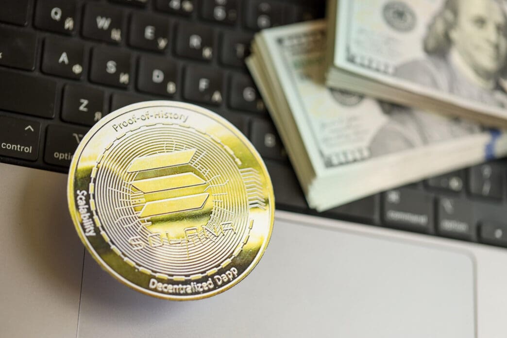 A physical gold Solana coin and a stack of US hundred-dollar bills rest on a laptop keyboard.