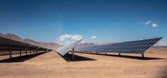 Expansive solar panel array stretching across a flat, arid desert landscape towards distant brown mountains under a bright blue sky.