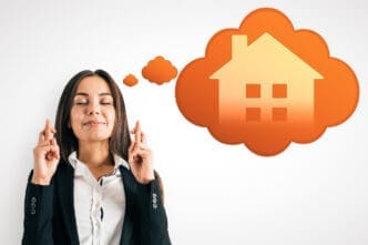 Woman with her eyes closed and fingers crossed, dreaming of a house visualized in an orange thought bubble.