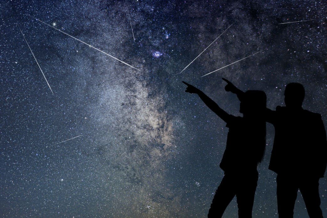 Silhouetted couple pointing up at the night sky filled with the Milky Way, numerous stars, and multiple bright streaking meteors.