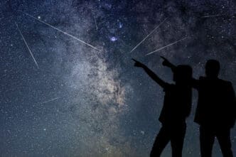Silhouetted couple pointing up at the night sky filled with the Milky Way, numerous stars, and multiple bright streaking meteors.