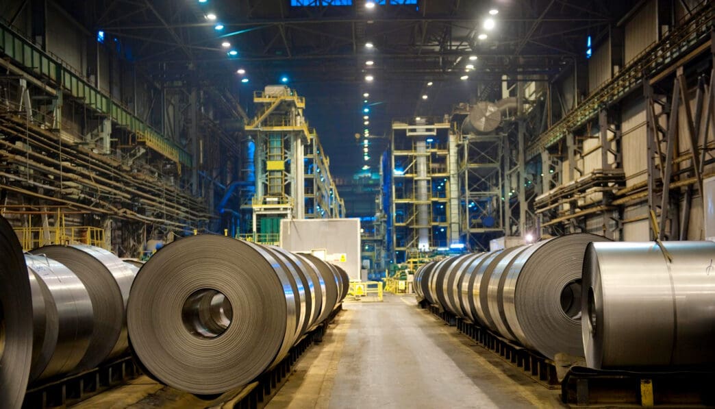 Massive coils of rolled steel lining an aisle inside a large, dimly lit industrial factory or mill.