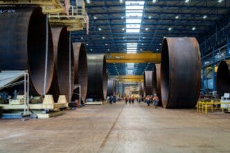 Enormous rolled steel sections for wind turbine towers lined up in a massive, brightly lit German factory hall with workers walking between them.