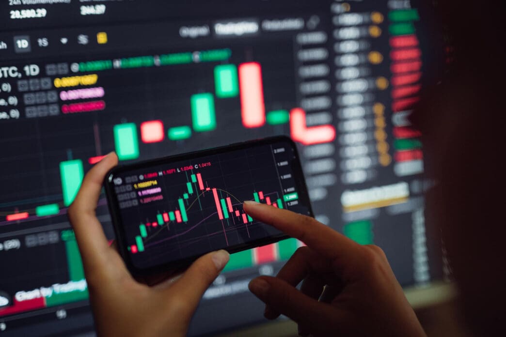 Hands holding a smartphone with a stock chart, a computer monitor with charts behind.