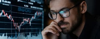 A stock market analyst wearing glasses studies a fluctuating chart on a computer screen.