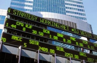 A dynamic stock market ticker board displaying glowing green text and financial data, set against the facade of a glass skyscraper