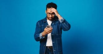 A stressed man in a denim jacket and glasses holds his smartphone to his face with one hand on his forehead against a blue background.