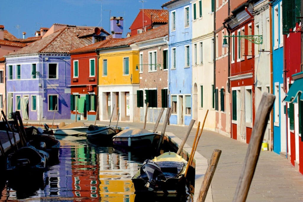 Small fishing boats moored on a narrow canal reflecting the brilliantly colored houses of Burano, Italy, under bright sunlight.