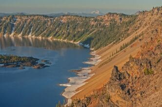 A high-angle view of Crater Lake at sunrise, showing Wizard Island and steep caldera walls.