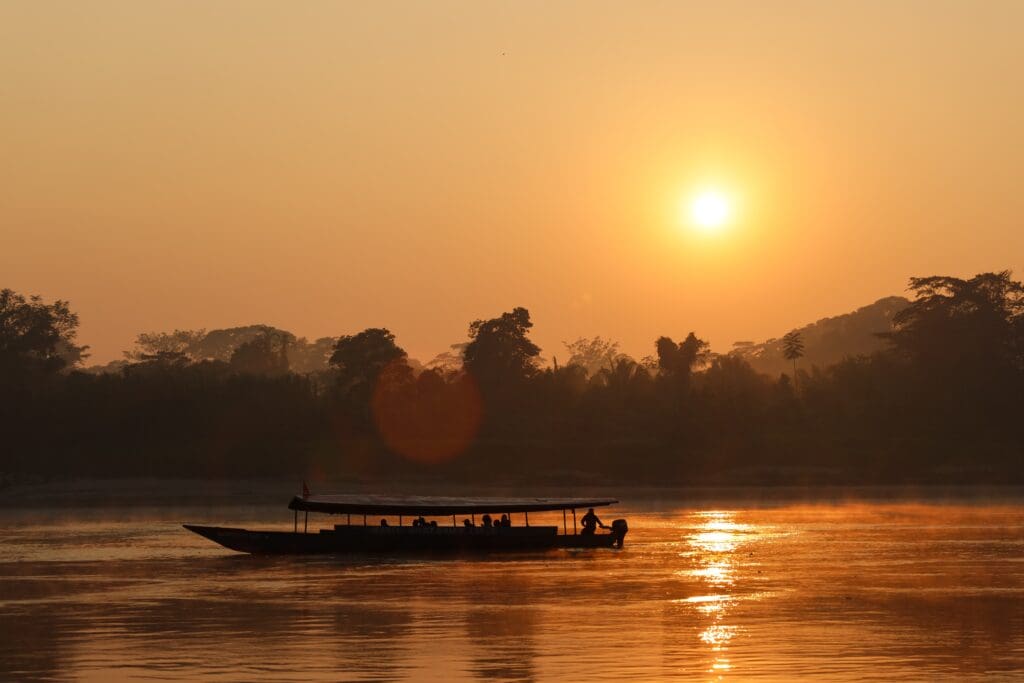 Silhouette of a long, covered river boat on a wide, misty river at sunrise, with the sky and water glowing orange and red.