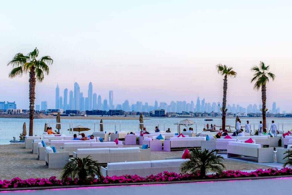 Beachfront dining area with white couches and palm trees overlooking the Dubai skyline at sunset.