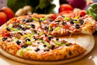 A slice of supreme pizza with melted cheese, olives, and green peppers being lifted from the whole pie on a wooden board