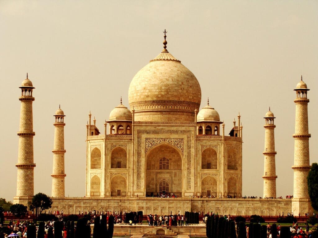Iconic Taj Mahal mausoleum, built from white marble, seen under a hazy sky with numerous visitors in the foreground.