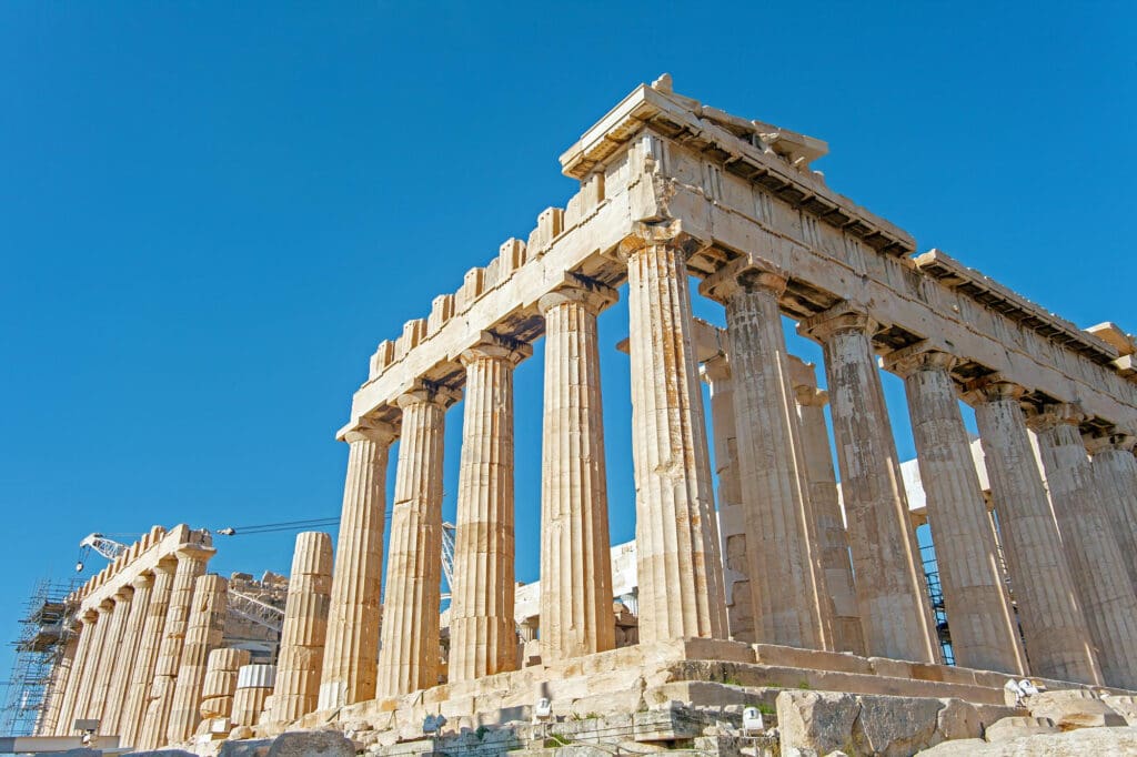A low-angle view of the ancient Parthenon temple in Athens, its massive marble columns standing against a bright blue sky.