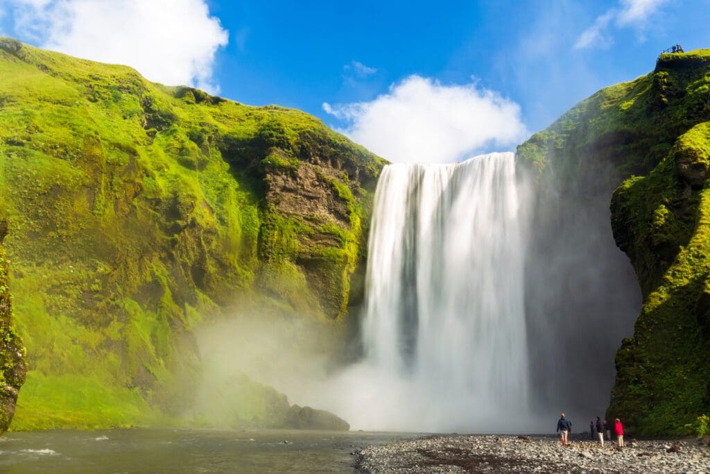 The massive Skógafoss waterfall in Iceland plunges over a mossy green cliff, with tiny people visible at its misty base.