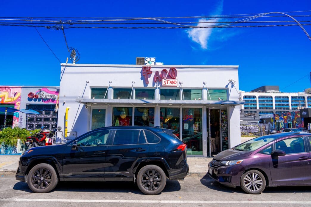 The exterior of "The Taco Stand" restaurant in Wynwood, Miami, with cars parked in front on a sunny day.