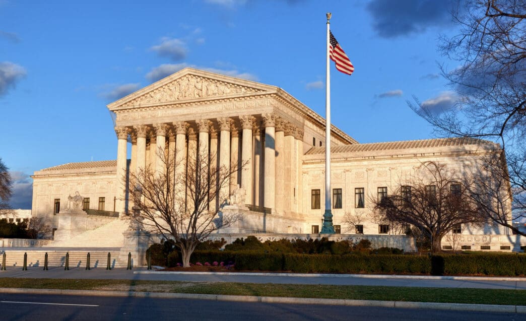 The US Supreme Court building, bathed in warm sunset light, with bare trees in the foreground.
