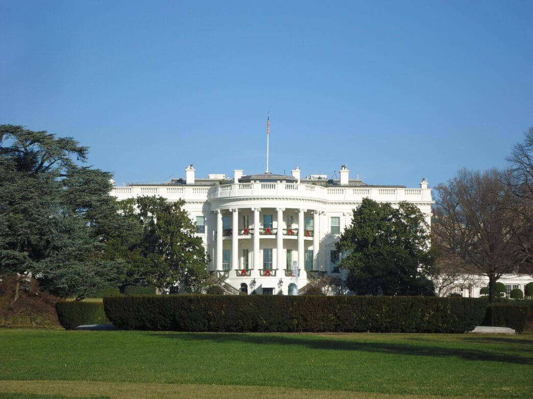 A straight-on view of the White House South Portico, with its curved balcony and columns, on a clear, sunny day.
