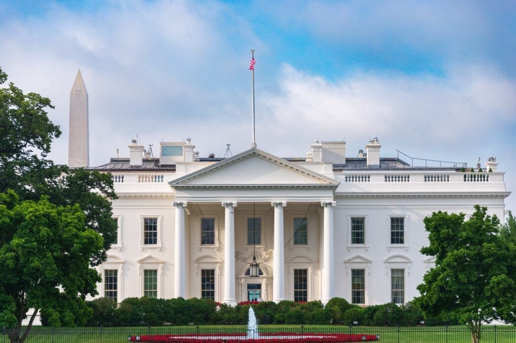 The White House, a grand white building with columns, stands under a blue sky with clouds, with the Washington Monument visible to the left.