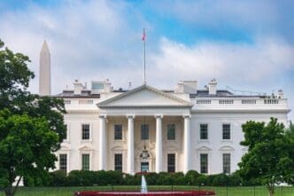 The White House, a grand white building with columns, stands under a blue sky with clouds, with the Washington Monument visible to the left.