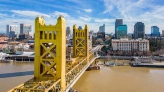 Aerial view of the bright yellow Tower Bridge over the muddy Sacramento River, with the city skyline behind it.