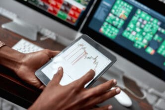 A trader's hands hold a tablet showing a stock chart, with monitors in the background.