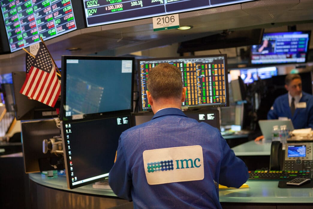 A stock trader with an IMC logo on his jacket works at a multi-monitor desk on the floor of the New York Stock Exchange.
