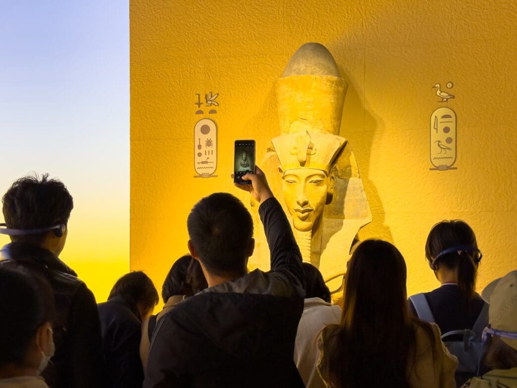 Museum visitors photographing a gold-colored pharaoh bust (Tutankhamun) mounted on a yellow wall with hieroglyphs