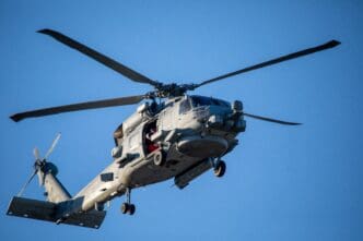 A U.S. Navy Sikorsky SH-60 Seahawk helicopter in light gray camouflage flies overhead against a clear blue sky.